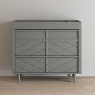 A modern gray Child Craft dresser with three drawers and geometric paneling stands on a light wood floor, perfectly paired with the F01792 - Universal Changing Table Topper in Lunar Gray for added convenience and style.