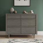 The Child Craft Surrey Hill 3 Drawer Dresser in Lunar Gray with angled diamond patterns stands against a green wall. On top are a ribbed vase, small round vase, toy train, and framed art above. A rug is seen on the floor.