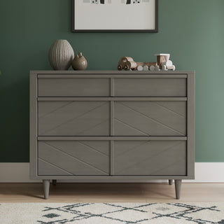 The Child Craft Surrey Hill 3 Drawer Dresser in Lunar Gray with angled diamond patterns stands against a green wall. On top are a ribbed vase, small round vase, toy train, and framed art above. A rug is seen on the floor.