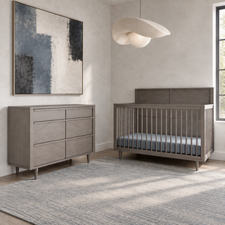 A Child Craft Surrey Hill 3 Drawer Dresser in Lunar Gray and a matching gray crib with horizontal slats in a room with abstract wall art and a pendant light.