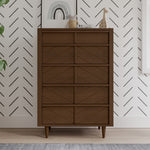 The Child Craft Surrey Hill 5 Drawer Chest in Toasted Chestnut stands against a white wall with a black chevron pattern. Decorative items, such as a giraffe figurine and framed art, sit on top, with a leafy plant partially visible on the left.