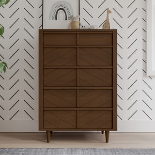 The Child Craft Surrey Hill 5 Drawer Chest in Toasted Chestnut stands against a white wall with a black chevron pattern. Decorative items, such as a giraffe figurine and framed art, sit on top, with a leafy plant partially visible on the left.