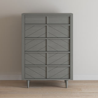 The Child Craft Surrey Hill 5 Drawer Chest in Lunar Gray features a geometric chevron front design, five drawers, and four tapered legs, shown against a white wall on a light wood floor.