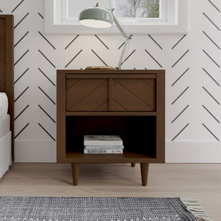 The Child Craft Surrey Hill Nightstand in Toasted Chestnut features angled legs and stands by a white wall with black chevron lines. It has a drawer, an open shelf with books, and a lamp on top. A bed, rug, and window are also visible.