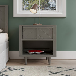 The Surrey Hill Nightstand by Child Craft in Lunar Gray stands beside a bed, featuring angled legs, a drawer, and an open shelf with two books. A bronze desk lamp sits on top and a window with a view of trees is above.