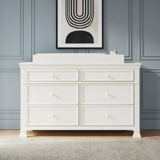 A Child Craft Westgate 3-Piece Nursery Set, featuring a white double dresser with six drawers, stands against a blue paneled wall and is topped with a changing pad and small container. A black and white abstract art print hangs above, completing this charming nursery ensemble.