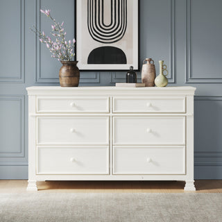 A Child Craft Westgate Double Dresser in Vintage Linen stands against a gray paneled wall, topped with vases and a pink-flowered branch. Black and beige abstract art hangs above, and a light rug covers the wooden floor.