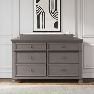 A gray double dresser with six drawers stands against a white paneled wall. A framed abstract art print hangs above, and a rectangular tray with small items sits atop the dresser—an elegant complement to the Child Craft Westgate 3-Piece Nursery Set with Crib, Dresser and Chest.