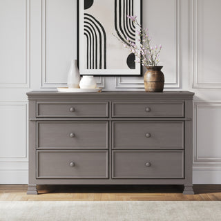 A Child Craft Westgate Double Dresser in Chelsea Gray sits against a white paneled wall, topped with ceramic vases, a bowl, and a brown vase with branches. Above it hangs abstract black-and-white artwork featuring curved lines.