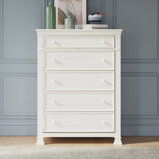 The Child Craft Westgate 5-Drawer Chest in Vintage Linen stands against a blue paneled wall, topped with green vases, books, and abstract art. This pine wood nursery dresser sits gracefully on a light wood floor.