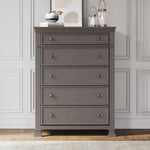 The Child Craft Westgate 5-Drawer Chest in Chelsea Gray stands against a white paneled wall, topped with a framed abstract art piece, decorative vase, and two glass lanterns. A light rug is partially visible on the hardwood floor.