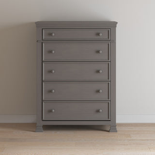 The Child Craft Westgate 5-Drawer Chest in Chelsea Gray stands against a light wall on a wooden floor, featuring five spacious drawers with round knobs and a tall, stylish gray wooden design.