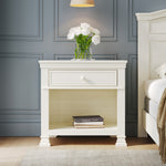 The Child Craft Westgate Nightstand in Vintage Linen, ideal for nurseries, features a drawer and open shelf styled with two books and a clear vase of white flowers, beside a bed against a blue paneled wall with a beige rug and wooden floor.