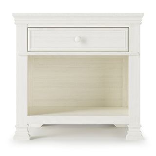 The Child Craft Westgate Nightstand in Vintage Linen is a white wooden nightstand with decorative legs, a single drawer with round knob, and an open shelf—ideal as nursery or baby room furniture thanks to its classic, simple design.