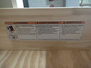 A warning label on the Child Craft Harmony 3-Drawer Dresser with Changing Table Topper, Brushed Cotton, in English, French, and Spanish advises securing the dresser to the wall and not placing TVs or a topper on it. Includes safety icons and instructions.