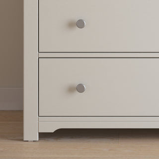 The Child Craft Harmony 3-Drawer Dresser with Changing Table Topper in Brushed Cotton features spacious drawers with round silver knobs, perfect for nursery storage. It stands on a wooden floor against a beige wall with white trim.