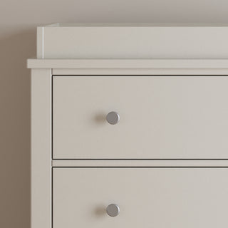 A close-up of the upper left corner of the Child Craft Harmony 3-Drawer Dresser with Changing Table Topper in Brushed Cotton shows two drawers with round metal knobs, set against a plain light-colored wall.
