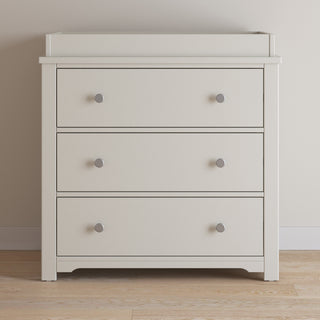 The Child Craft Harmony 3-Drawer Dresser with Changing Table Topper in Brushed Cotton offers stylish nursery storage, featuring a cream finish and silver knobs—perfect against a light beige wall on a wooden floor.