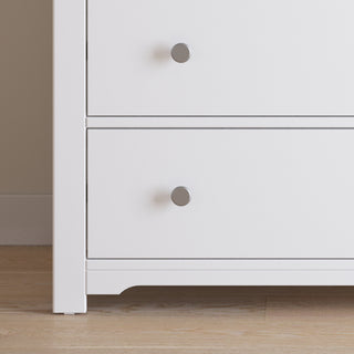 Close-up of the Child Craft Harmony 3-Drawer Dresser with Changing Table Topper in Matte White, featuring round silver knobs. Perfect for nursery storage, shown on a light wood floor beside a beige wall with white baseboards.
