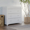 The Child Craft Harmony 3-Drawer Dresser with Changing Table Topper in matte white stands on a wood floor beside woven baskets and a potted plant, with light blue patterned wallpaper creating a bright, stylish nursery scene.