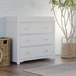 The Child Craft Harmony 3-Drawer Dresser with Changing Table Topper in matte white stands on a wood floor beside woven baskets and a potted plant, with light blue patterned wallpaper creating a bright, stylish nursery scene.