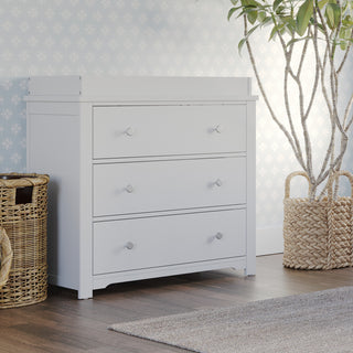 The Child Craft Harmony 3-Drawer Dresser with Changing Table Topper in matte white stands on a wood floor beside woven baskets and a potted plant, with light blue patterned wallpaper creating a bright, stylish nursery scene.