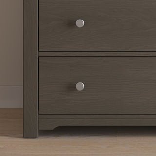 A close-up of the lower section of the Child Craft Harmony 3-Drawer Dresser with Changing Table Topper in Dapper Gray, featuring round silver knobs. The dresser stands on a light wood floor beside a beige wall.