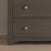 A close-up of the lower section of the Child Craft Harmony 3-Drawer Dresser with Changing Table Topper in Dapper Gray, featuring round silver knobs. The dresser stands on a light wood floor beside a beige wall.