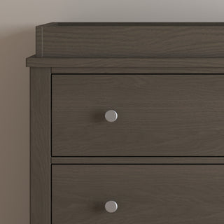 A close-up of the upper left corner of the Child Craft Harmony 3-Drawer Dresser with Changing Table Topper in Dapper Gray, featuring two drawers with round silver knobs, set against a plain light-colored wall.