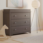 A modern nursery with the Child Craft Harmony 3-Drawer Dresser with Changing Table Topper in Dapper Gray, a white tripod floor lamp, a light-toned rocking horse, a beige rug on wood flooring, and minimalist artwork above the dresser.