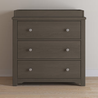 The Child Craft Harmony 3-Drawer Dresser with Changing Table Topper in Dapper Gray stands on a light wood floor against an off-white wall, featuring silver round knobs for a modern touch.