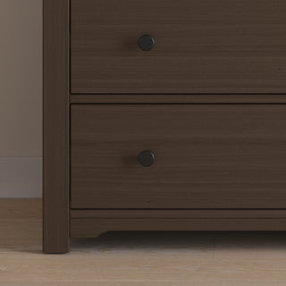 A close-up of the Harmony 3-Drawer Dresser with Changing Table Topper by Child Craft in Brushed Truffle, showing two drawers with round black knobs. The dresser sits on a light wooden floor beside a beige wall.