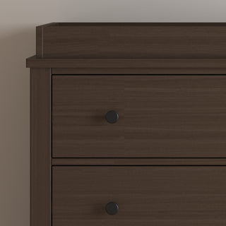 A close-up of the upper section of the Child Craft Harmony 3-Drawer Dresser with Changing Table Topper in Brushed Truffle shows two drawers with round black knobs, set against a plain light wall.
