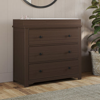 The Child Craft Harmony 3-Drawer Dresser with Changing Table Topper in Brushed Truffle stands on a wooden floor beside a woven basket with a green plant; above it are a round mirror and wall decor.