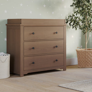 The Child Craft Harmony 3-Drawer Dresser with Changing Table Topper in Dusty Heather stands on a light wood floor, set against light green star-patterned wallpaper, with a leafy plant, woven basket, and white textured bin nearby.