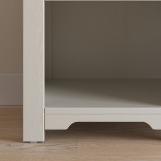 A close-up of the bottom section of the Child Craft Harmony Nightstand in Brushed Cotton on a wood floor against a beige wall. The empty nightstand features a small decorative cutout at its base.