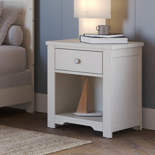 The Harmony Nightstand by Child Craft in Brushed Cotton features a drawer and open shelf, holding a lamp, two books, and a mug on top, with a wooden decor piece below. It sits beside a bed with light bedding.