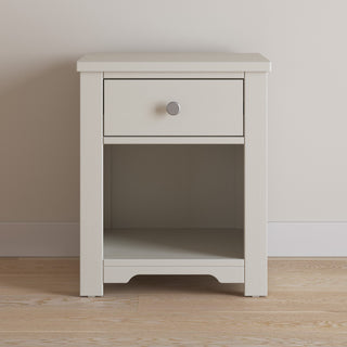The Child Craft Harmony Nightstand in Brushed Cotton is a simple, light-colored wood piece with one drawer, a round metal knob, and an open shelf below, shown on a wooden floor against a plain wall.
