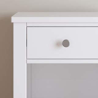 A close-up of the upper left corner of the Child Craft Harmony Nightstand, Matte White, featuring a single drawer with a round silver knob, set against a plain light wall and crafted with child-safe finishes.