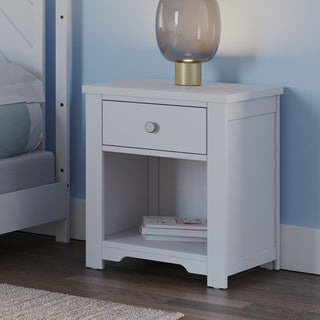 The Child Craft Harmony Nightstand in Matte White offers child-safe finishes, a drawer, and an open shelf for storage. Topped with a modern glass lamp with a gold base, it complements the serene bedside setting.