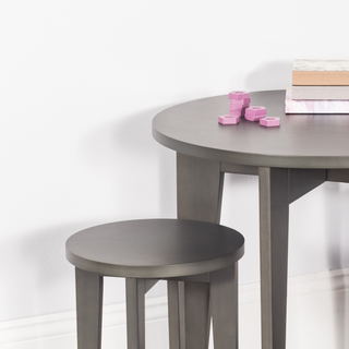 The Child Craft Geo Table and Stools Set in Dapper Gray features a round wooden table with a matching stool, three stacked books, and several small pink hexagonal pieces arranged on the tabletop against a white wall.