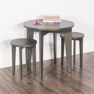 The Child Craft Geo Table and Stools Set in Dapper Gray features a small round table with two matching stools on a wooden floor. Stacked books and pink wooden blocks sit atop the table, set against a white wall with baseboard.
