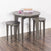 The Child Craft Geo Table and Stools Set in Dapper Gray features a small round table with two matching stools on a wooden floor. Stacked books and pink wooden blocks sit atop the table, set against a white wall with baseboard.