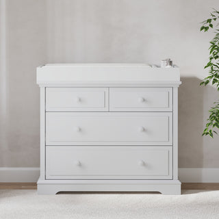The Child Craft F01792 Universal Changing Table Topper in Matte White sits on a changing table with two small top drawers and two larger bottom drawers, placed against a light wall on carpet. A leafy green plant is partly visible to the right.