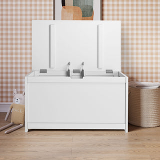 The Child Craft Harmony 33" Kids Toy Box in matte white sits open on a wooden floor, displaying spacious storage. A stuffed animal is on the left, a woven basket on the right, with checked wallpaper and framed abstract art in the background.