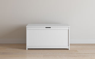The Child Craft Harmony 33" Kids Toy Box in matte white, featuring a flat lid and small handle, sits on a wooden floor against a plain, light-colored wall.