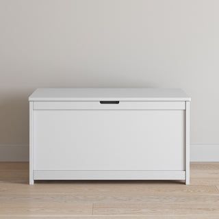 The Child Craft Harmony 33" Kids Toy Box in matte white with a hinged lid is placed against a beige wall on a light wood floor.