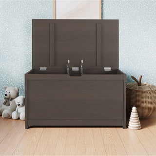 The Child Craft Harmony 33" Kids Toy Box in Dapper Gray sits open on a light wood floor, with two stuffed bears, a woven basket, and a stacked wooden toy nearby. Light blue wallpaper with a white leaf pattern decorates the background.