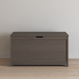 The Child Craft Harmony 33" Kids Toy Box in Dapper Gray sits on a light wood floor by a plain beige wall. It features a simple design with a small handle cutout on the front panel.