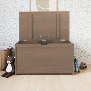 The Child Craft Harmony 33" Kids Toy Box in Dusty Heather sits against a wall with leafy wallpaper. A brown teddy bear is on the left, while books and a toy rest on the right, all atop a light wood floor.